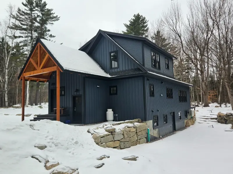 Side view of a blue custom home featuring stone wall landscape and winter woodland backdrop